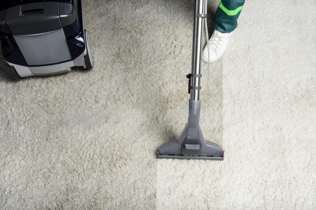 A person uses a carpet cleaner on a light-colored carpet, showing a noticeable contrast between the cleaned and uncleaned areas.