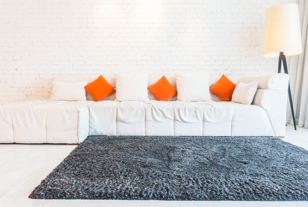 A spacious white sofa with four orange and three white pillows stands against a white brick wall. A tall floor lamp is on the right. A dark gray rug covers the light-colored floor.