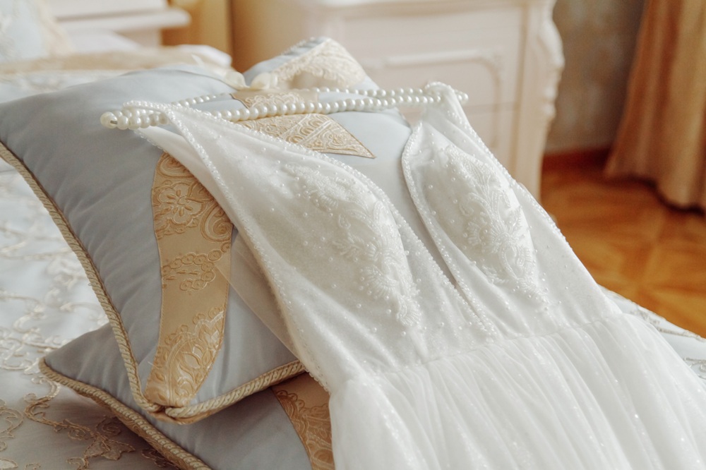 A white lace wedding dress and pearl necklace are displayed on pillows atop a bed in a softly lit room.