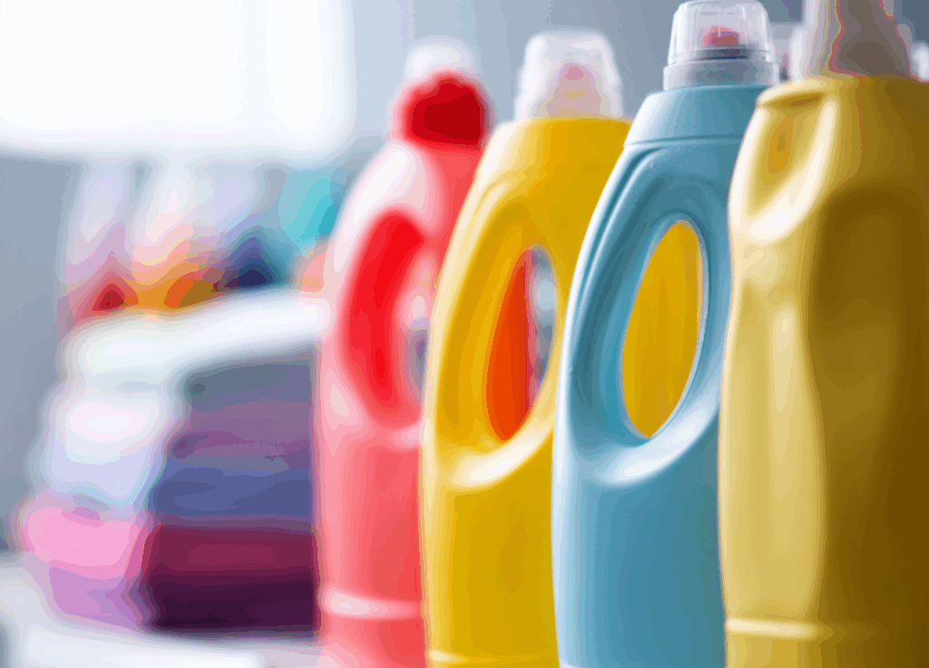 Colorful plastic detergent bottles are lined up in the foreground, with folded laundry stacks visible in the blurred background.