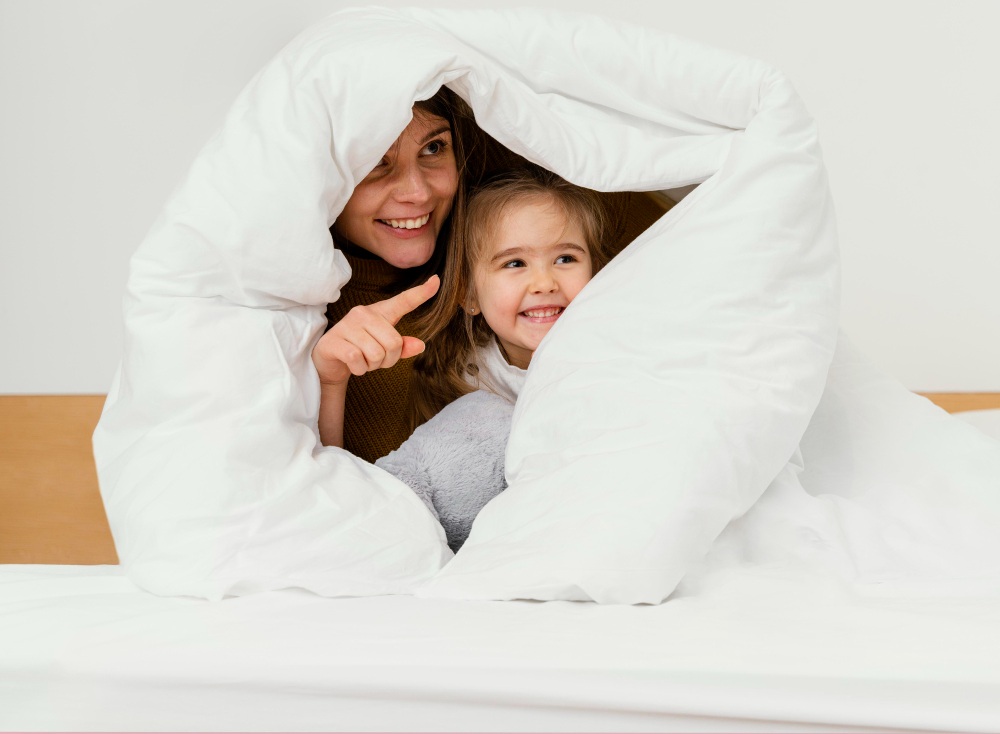 An adult and a child smile together under a white blanket on a bed; the freshly laundered comforter adds to their cozy moment as the adult points at something off camera.