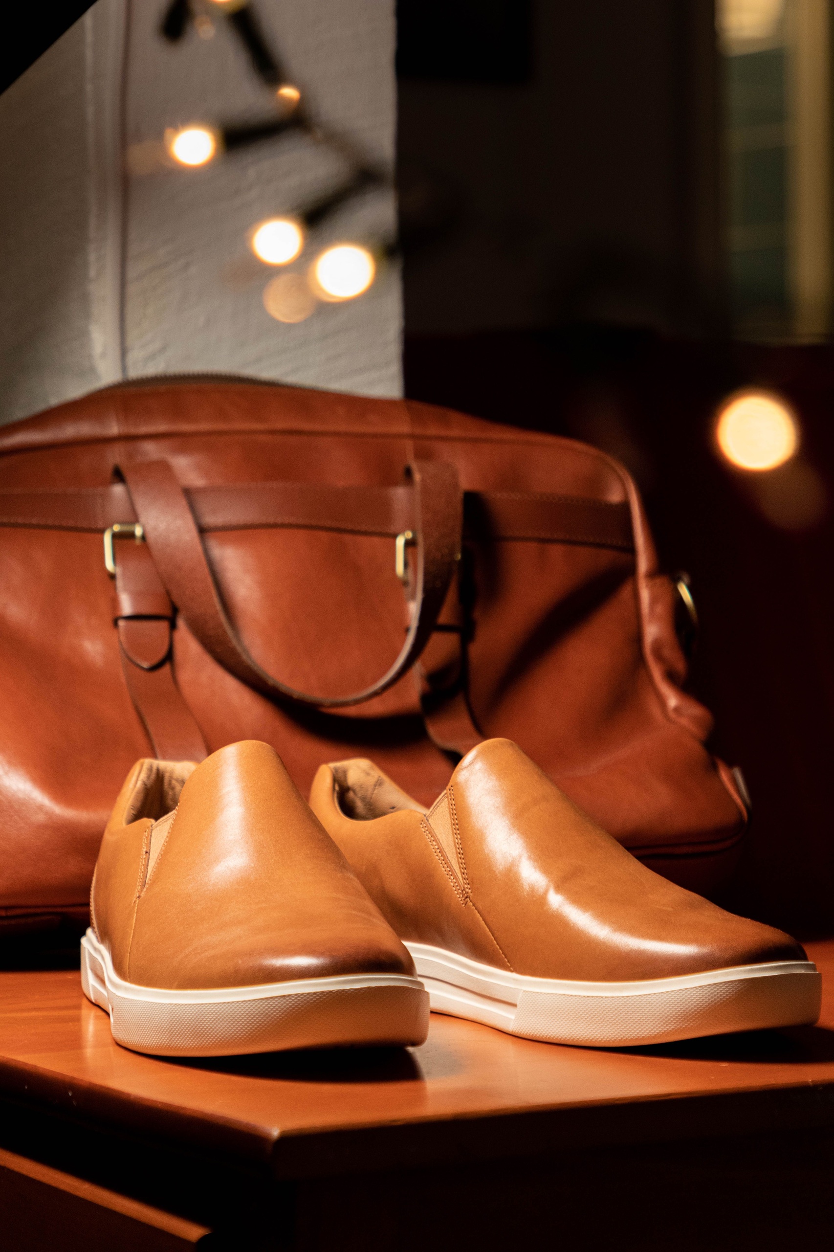 A pair of tan slip-on shoes with white soles is displayed on a wooden surface in front of a matching brown leather bag.