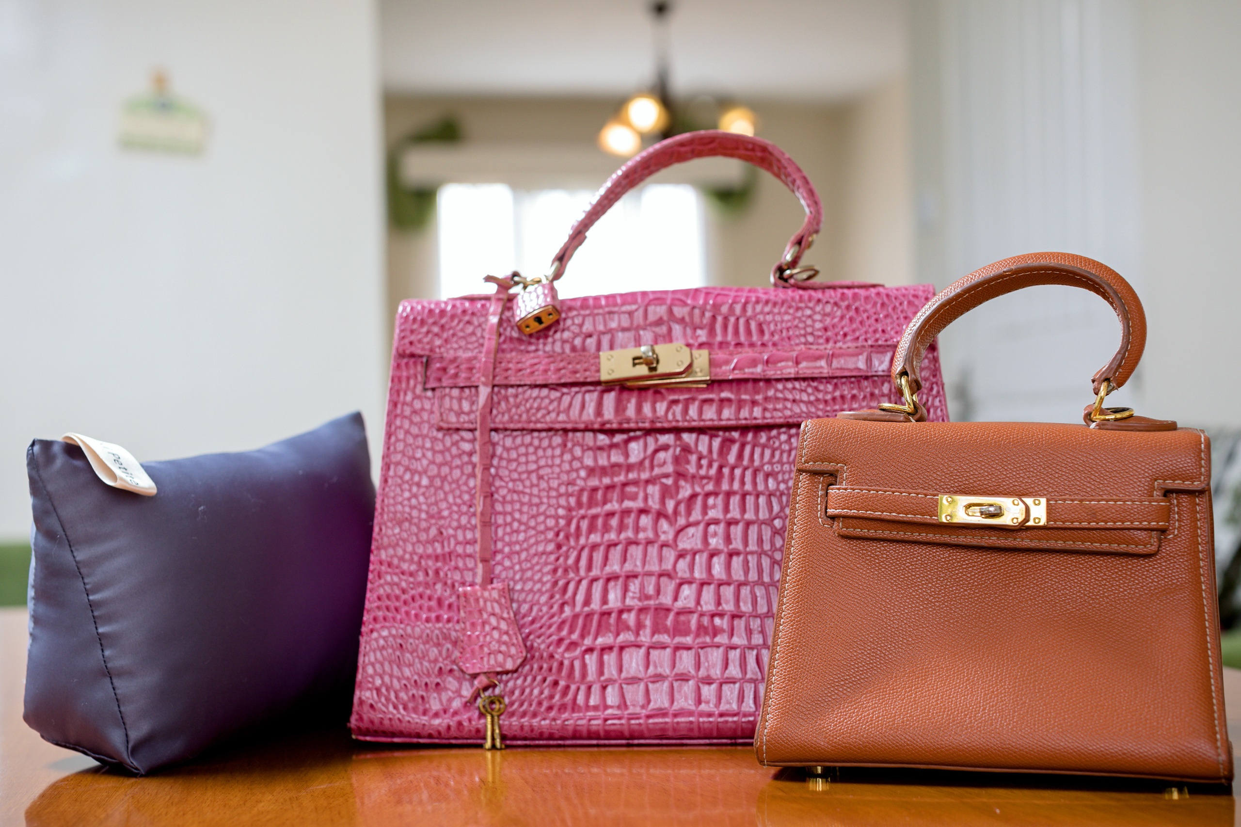 A purple pillow sits next to a large pink crocodile leather handbag and a small brown leather handbag on a wooden table.
