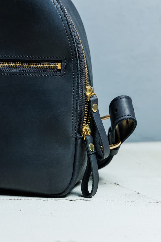 Close-up of a black leather bag with a gold zipper, a front pocket, and a strap with metal hardware against a light gray background.