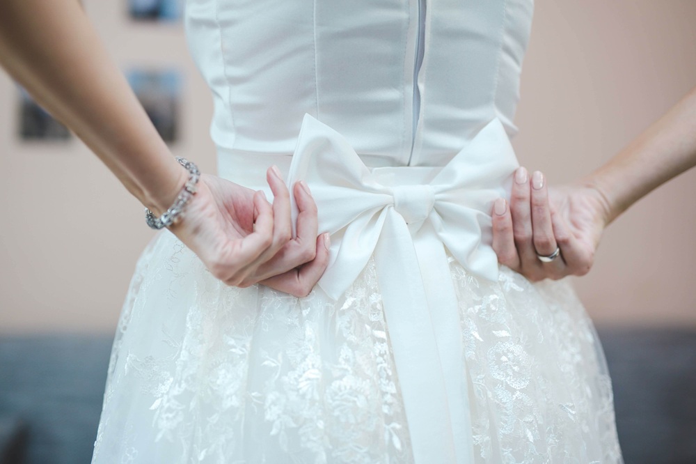 A person ties a large white bow on the back of a lace wedding dress, focusing on the hands and dress details.
