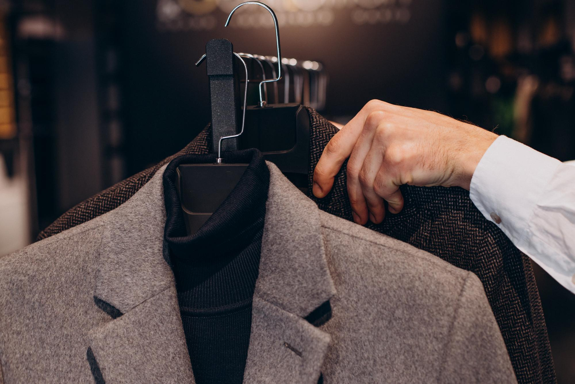 A person’s hand holding a gray blazer and a dark turtleneck sweater on a hanger in a clothing store.