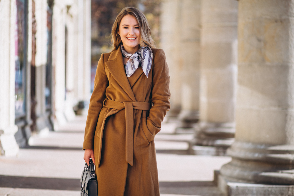 A woman wearing a brown coat and patterned scarf stands outdoors, smiling, with one hand in her pocket and holding a bag in the other. Stone columns line the background.